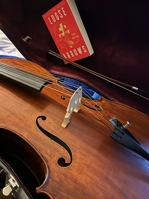 A cello in its case with the Lynn Horton novel Loose Arrows stuck to the interior lid of the case